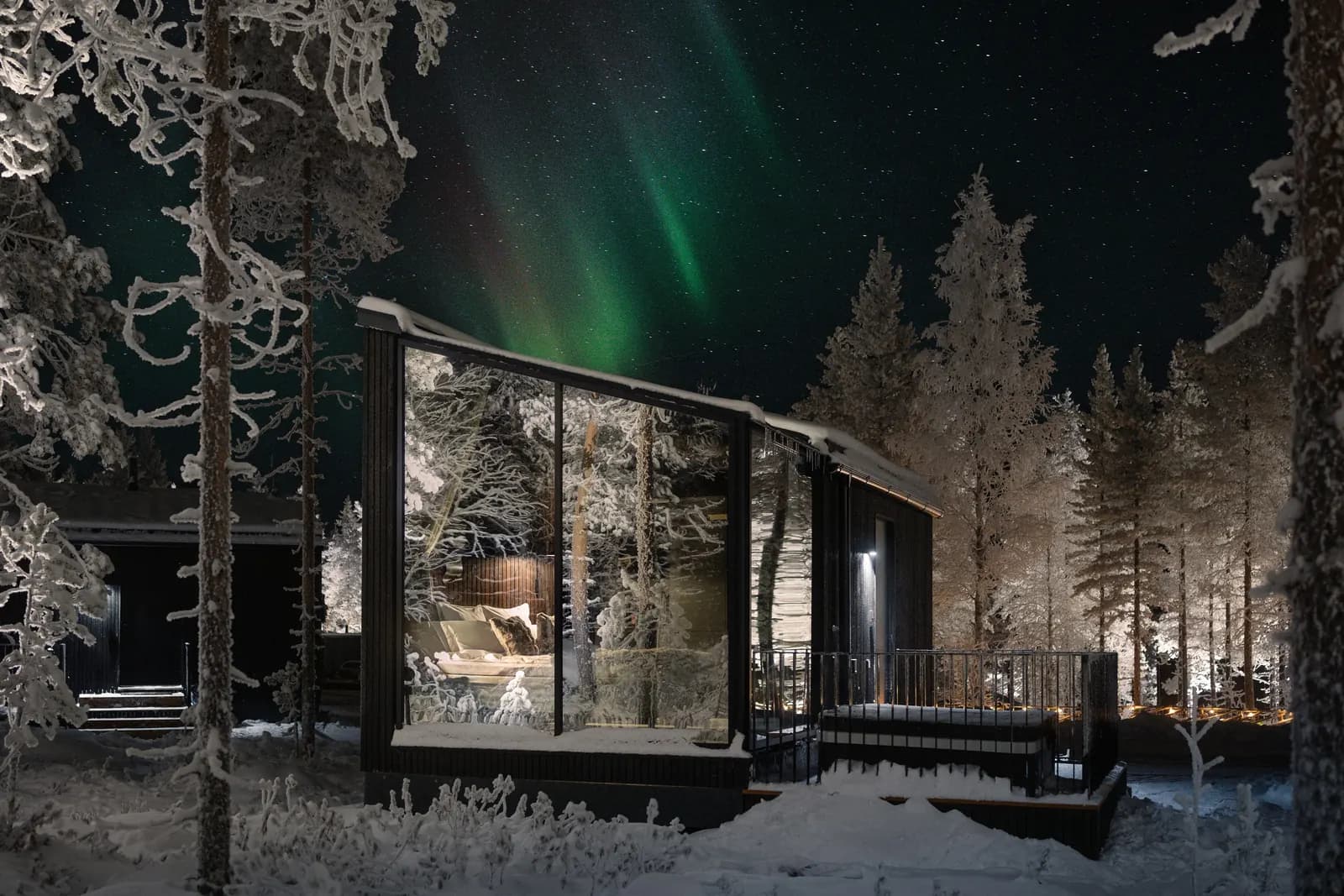 A Halo Hillside glass igloo lit up at night under green Northern Lights, among snow-covered trees
